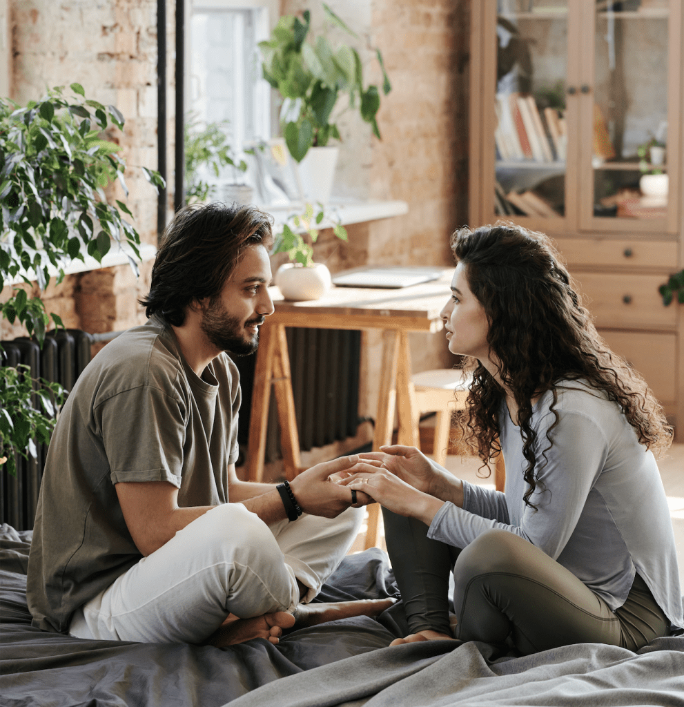 A man and woman sit on a bed facing each other and holding hands, illustrating that Susan Gonzales provides tips for healthy communication in relationships.